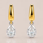 A pair of gold and silver drop earrings, each featuring a polished gold teardrop-shaped top and a pear-cut clear gemstone in a silver bezel setting below, shown on a light beige background.