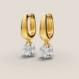 Gold hoop earrings with heart-shaped diamond drops on a light beige background