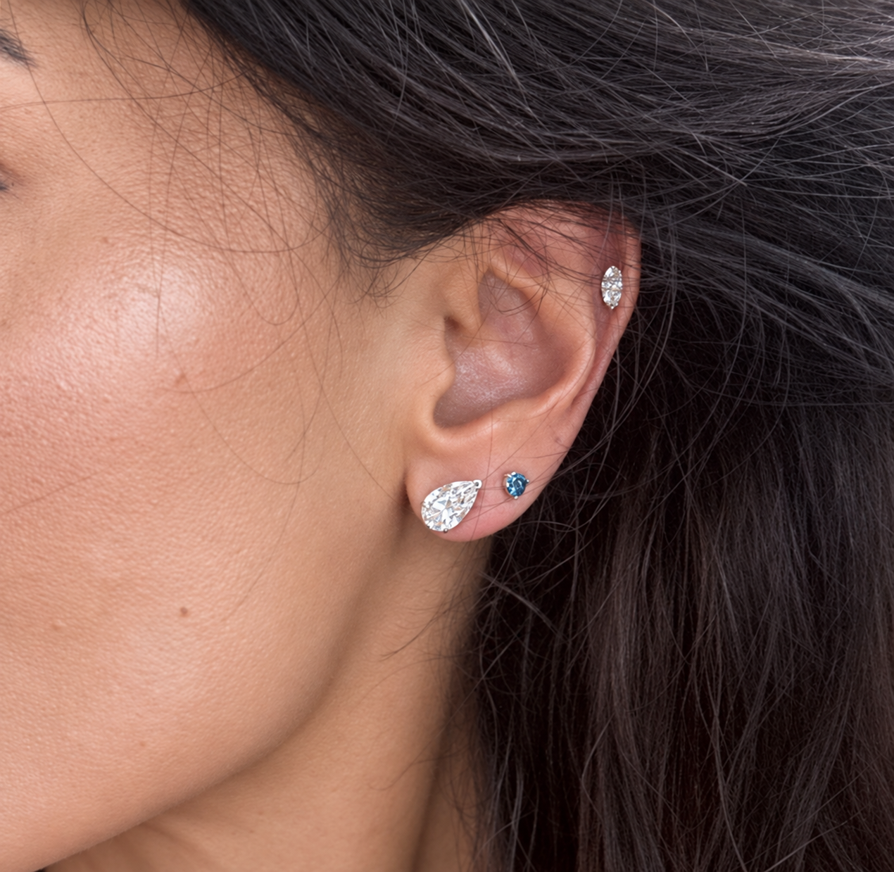Close-up of a person's ear wearing multiple earrings, including a teardrop-shaped silver stud with a clear gemstone, a small blue gemstone stud, and a silver hoop earring.