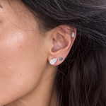 Close-up of a person's ear wearing multiple earrings, including a teardrop-shaped silver stud with a clear gemstone, a small blue gemstone stud, and a silver hoop earring.