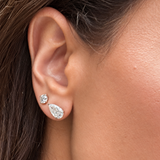 Close-up of a person's ear wearing two silver-tone stud earrings, one small round stud and one larger teardrop-shaped stud, both set with clear gemstones.