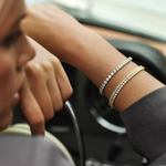A woman's arm showing a tennis bracelet with multiple diamond charms, in a car interior.