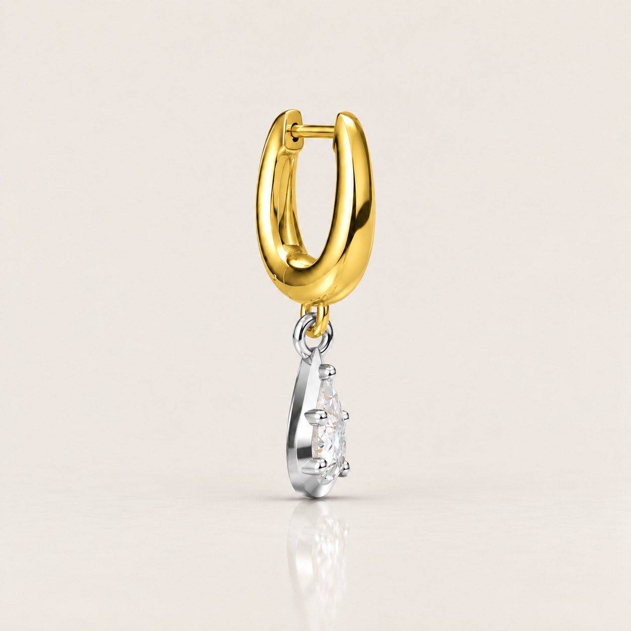 A gold huggie hoop earring with a dangling silver-toned charm featuring a clear gemstone, shown on a light beige background with a soft reflection below.