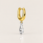 A gold huggie hoop earring with a dangling silver-toned charm featuring a clear gemstone, shown on a light beige background with a soft reflection below.