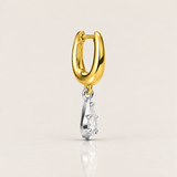 A gold huggie hoop earring with a dangling silver-toned charm featuring a clear gemstone, shown on a light beige background with a soft reflection below.