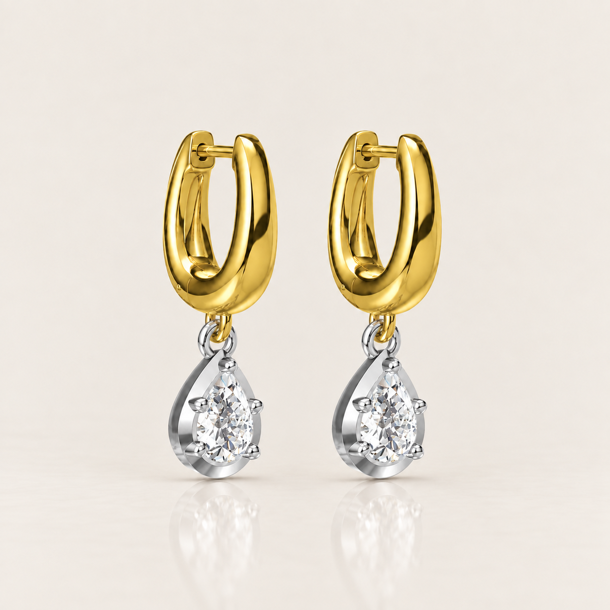 A pair of gold and silver drop earrings, featuring a polished yellow gold huggie hoop with a dangling pear-cut clear gemstone in a silver setting, shown on a light beige background.