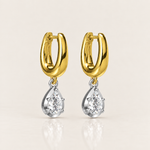 A pair of gold and silver drop earrings, featuring a polished yellow gold huggie hoop with a dangling pear-cut clear gemstone in a silver setting, shown on a light beige background.