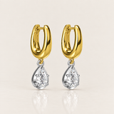 A pair of gold and silver drop earrings, featuring a polished yellow gold huggie hoop with a dangling pear-cut clear gemstone in a silver setting, shown on a light beige background.